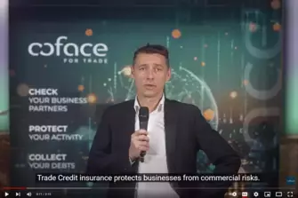 Sie sehen hier einen Screenshot eines Youtube-Videos. Dieser zeigte den Coface-Experten Frederic Orillard mit einem Mikrofon in der Hand, während er vor einem Banner mit Coface-Logo steht. Er trägt einen schwarzen Anzug und ein weißes Hemd darunter. Er steht frontal zur Kamera und wird von vorne angeleuchtet.
