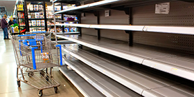 In een supermarkt staat een leeg winkelwagentje voor een leeg schap. Op de achtergrond zijn twee mensen te zien